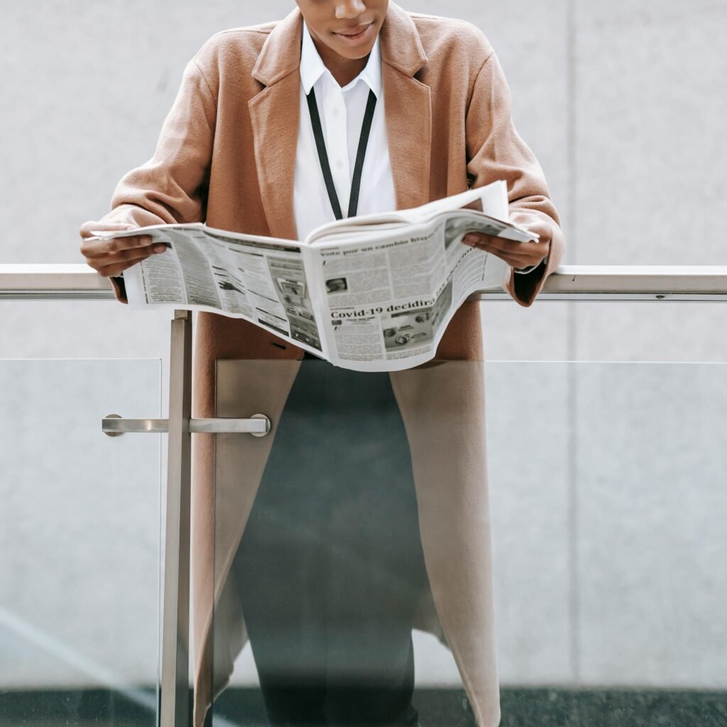 Crop content African American businesswoman in formal clothes reading fresh newspaper and leaning on glass railing of contemporary office building